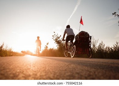 Man And Woman Are On The Bikes On The Road Trip With The Baby Coach. Strong Warm Sunset Light. Wide View From Below. Back Side Shot.