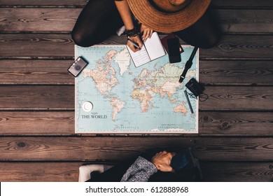 Man And Woman Planning Vacation Using A World Map And Other Travel Accessories. Woman Noting The  Discussion Points In A Small Diary.