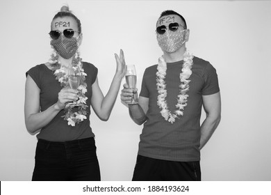 man and woman posing near white wall with glasses of champagne in hand. boy and girl wearing hawaiian flowers and face masks to protect from coronavirus. celebrating new year 2021 at home quarantine.