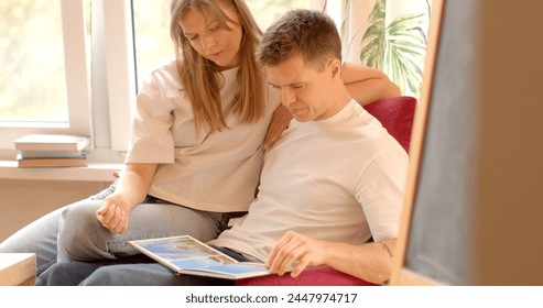 Man and woman reading book. Warm and inviting ambiance of nursery, young parents sit side by side, immersed in nostalgic journey of flipping through a photo album. Use printed publications on paper.  - Powered by Shutterstock