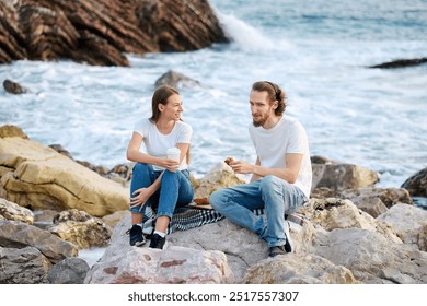 A man and woman relaxing on coastal rocks with snacks, facing each other and the ocean. - Powered by Shutterstock