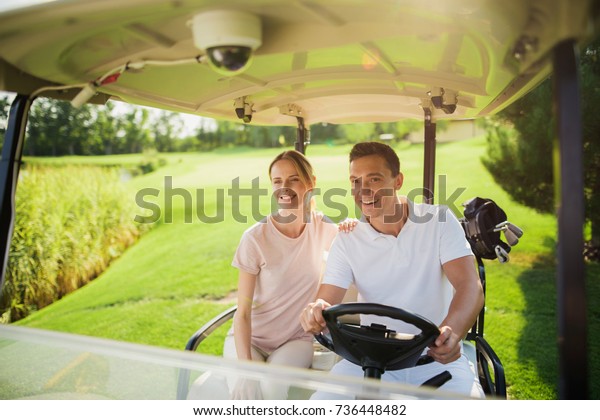 A man and a woman ride a white golf cart on the
golf course. A man drives a car, a woman sits next to him, putting
her hand on his shoulder