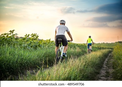 Man And Woman Riding Bicycles In A Field At Sunset In The Fall, Summer. The Girl And The Guy On The Bike Involved In Cycling. Walk On Bicycles Outdoors.