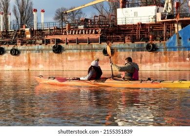 Man And Woman (romantic Couple) Paddles A Kayak On The Evening Winter River Near Old Rusty Ship