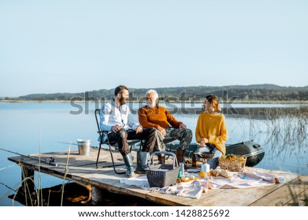 Man and woman with senior grandfather having a picnic with vegetables and fresh caught fish on the lake in the morning