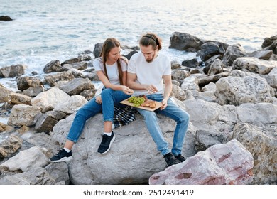 A man and woman share a meal on the rocky shore beside calm sea waters in evening light. - Powered by Shutterstock