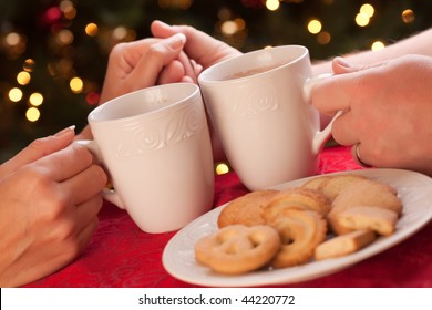 Man And Woman Sharing Hot Chocolate And Cookies In Front Of Holiday Lights.