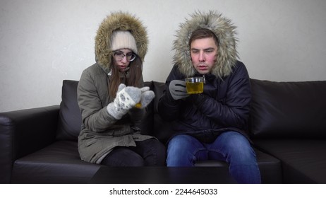 A man and a woman sit at home in jackets and drink tea. The house is very cold because of the prohibition to raise the temperature in the house. A young couple sits without heating. - Shutterstock ID 2244645033
