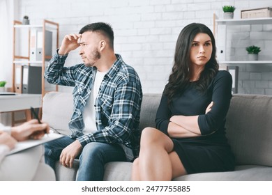 A man and a woman sit on a couch in a therapy session. The man is looking down, with his hand on his forehead, while the woman is sitting with her arms crossed, looking straight ahead - Powered by Shutterstock