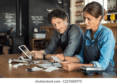 Man and woman sitting in cafeteria discussing finance for the month. Stressed couple looking at bills sitting in restaurant wearing apron. Café staff sitting together looking at expenses and bills. - Powered by Shutterstock