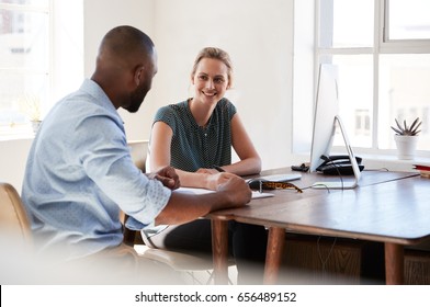 Man And Woman Sitting At A Desk Talking In An Office Smiling