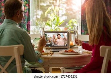 Man and woman sitting at the dining table, having dinner, drinking Champagne and having video call with senior parents on laptop. Staying home, quarantine and social distancing celebration of event. 