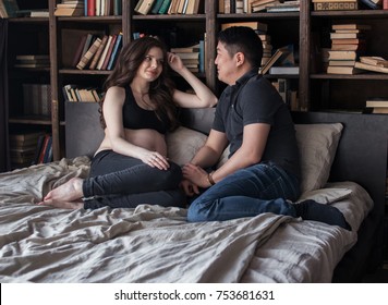 man and woman sitting on the bed, woman pregnant, looking at each other couple smiling. happiness. joy. pregnancy. bookcase - Shutterstock ID 753681631