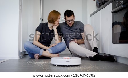 man and woman are sitting on a floor in home and trying to switch on a robotic vacuum cleaner using app in smartphone, examining modern gadgets Stock photo © 