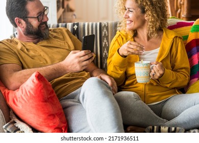 Man And Woman Smile And Enjoy Leisure Activity At Home In Morning Breakfast Time Together. People Sitting On The Sofa In Apartment For Real Life. Couple Use Phone And Drink Tea Having Fun On The Couch