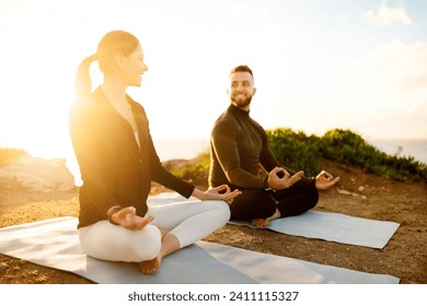 Man and woman smiling, sitting in lotus positions on yoga mats, meditating in the serene light of sunset on cliff overlooking the ocean - Powered by Shutterstock