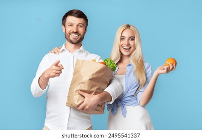 A man and a woman stand together against a blue background. The man is holding a brown paper bag filled with groceries and pointing to the side with his right hand - Powered by Shutterstock