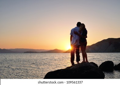 Man and woman standing arm in arm on a rock by the sea and watching the sunset