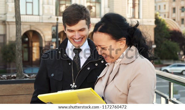 A man\
and a woman are standing in the middle of the street. A woman is\
holding a bright yellow folder with documents in her hands. They\
look at it and discuss something. Then people\
laugh