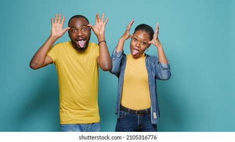 Man And Woman Sticking Tongue Out And Fooling Around In Front Of Camera. Playful People Doing Funny And Comic Faces, Enjoying Goofy Joke Together In Studio. Couple Showing Childish Behavior.