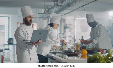 Man And Woman Using Laptop To Cook Food Recipe In Kitchen, Making Gourmet Dish. Team Of People Cooking Professional Authentic Meal With Computer Online Website, Preparing Ingredients.