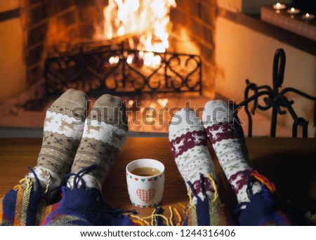 Man and woman in warm knitted socks with cups of hot punch in front of the fireplace