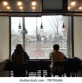 A Man And A Woman Are Watching The Snow Falling Outside Window At A Cafe. 