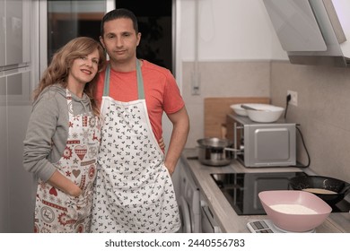 A man and woman wearing aprons standing in a modern kitchen, smiling with cooking utensils and a bowl of flour nearby.
