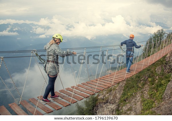 A man and a woman wearing a safety net. They\
are walking on a rope bridge over an abyss. Crossing a suspension\
bridge in the mountains. Extreme sport. The view from the back. On\
a background of clouds