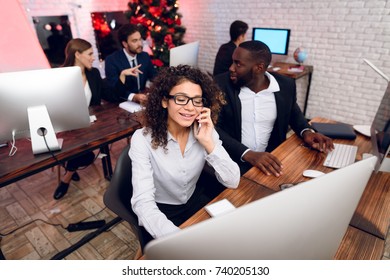 A Man And A Woman Are Working In The Office On The Eve Of The New Year. They Are Posing In Their Workplaces And Are Busy With Work. Behind Them Sit Their Colleagues. In The Room A Christmas Tree.