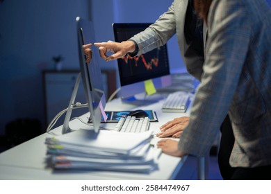 Um homem e uma mulher estão trabalhando juntos em um computador. A mulher está apontando para a tela do computador, e o homem está olhando para ela. A tela do computador mostra um gráfico com uma tendência de queda