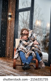 Man And Woman Wrapped In Blanket Cuddling In Chair Outside Scandinavian House Barnhouse. Couple In Love Hugging Outdoors Near Building With Panoramic Window Under Winter Snow.