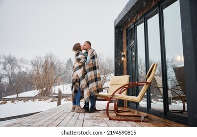 Man And Woman Wrapped In Blanket Kissing Outdoors Under Winter Snow. Happy Couple Sharing Romantic Moment While Standing Near Chairs Outside Scandinavian House Barnhouse With Panoramic Windows.