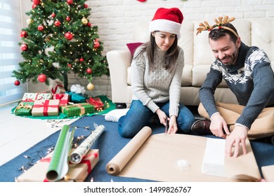 Man and woman wrapping paper together over gift box while sitting against Christmas tree at home - Powered by Shutterstock