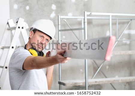 man work,  checks the straightedge to level the wall, with ladder and scaffolding in the interior construction site on background