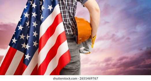 man in work clothes with hard hat and American flag in hand standing against sunset background. labor day concept