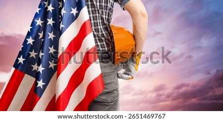 man in work clothes with hard hat and American flag in hand standing against sunset background. labor day concept