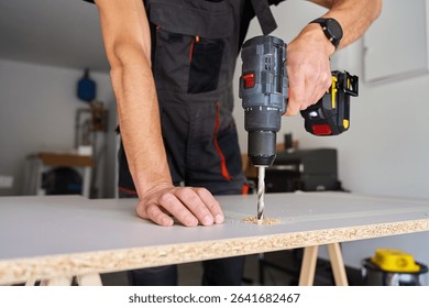 Man in work overalls bib using cordless power drill to make hole in laminated wood board. Male carpenter working in home workshop, drilling chipboard on workbench