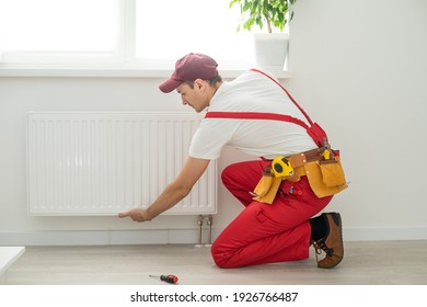 Man in work overalls using wrench while installing heating radiator in room. Young plumber installing heating system in apartment. Concept of radiator installation, plumbing works and home renovation