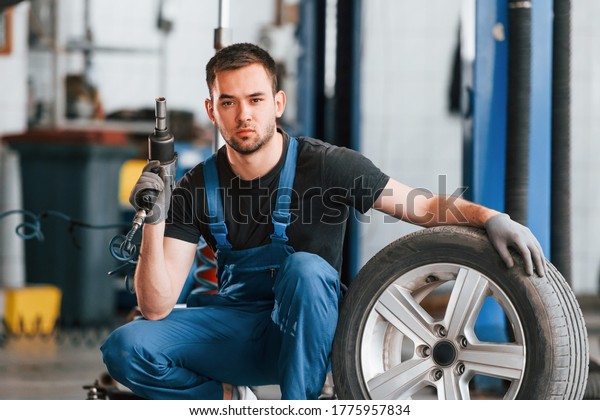 Man in work uniform sitting with car\
wheel indoors. Conception of automobile\
service.
