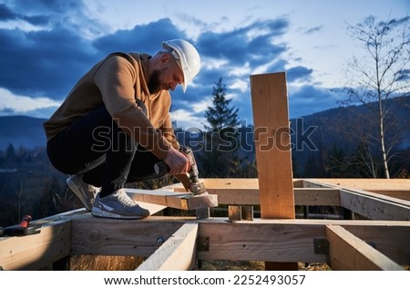 Man worker building wooden frame house on pile foundation. Carpenter installing furniture for wooden joist, using hammer and screwdriver. Carpentry concept.