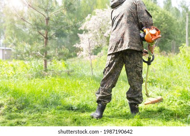 A Man Worker In Camo Workwear Mowing Tall Grass With Petrol Lawn Trimmer Or Lawn Mower In City Park, Garden Or Backyard. Gardening Care Tools And Equipment. Process Of Lawn Trimming With Hand Mower.