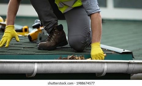 A man worker is cleaning a clogged roof gutter from dirt, debris and fallen leaves to prevent water and let rainwater drain properly.