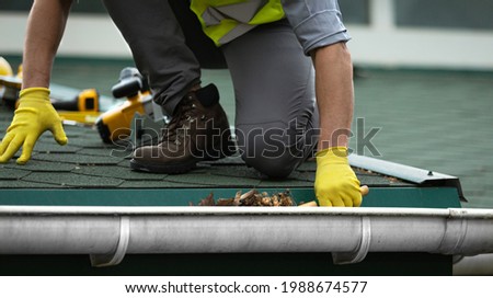 A man worker is cleaning a clogged roof gutter from dirt, debris and fallen leaves to prevent water and let rainwater drain properly.