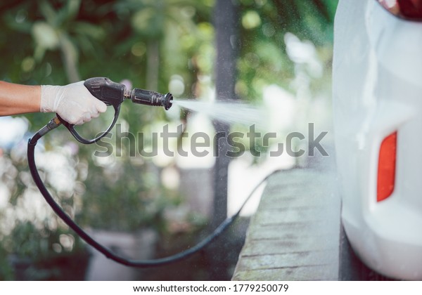Man worker in garage using a high pressurised foam\
injector spraying on a dirty white car. Car washing and car care\
concept in rainy season. Manual labor spray a liquid soap on a\
dirty car.