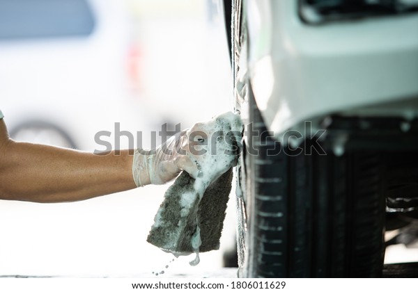 Man worker in garage washing a dirty car\
by using a car washer soap and a brush to wash a dirt from a tires.\
Car washing service concept with\
copyspace.