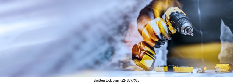 Man worker holds an electric screwdriver in his hands close-up against the background of a construction tool and a concrete wall. Long banner with glow effects - Powered by Shutterstock