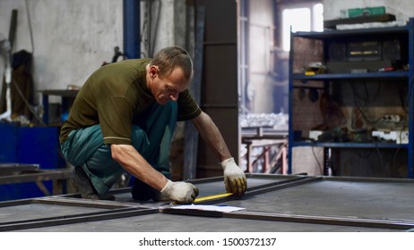 Man Worker Locksmith Measures Tape Measure Length Of Metal Bars. He Makes Marks With Pencil In Workshop. He Is In Uniform And Gloves In His Workplace. Work With Metal In Production.