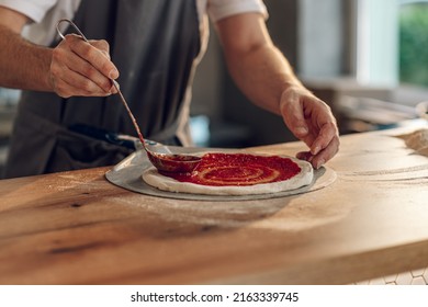 Man worker in a pizza place placing ingredients on pizza base. Special sauce is applied on the pizza dough and pizza ingredients are placed on it. - Powered by Shutterstock