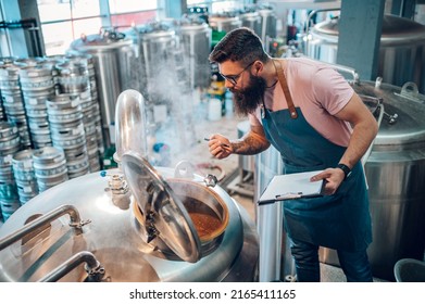 Man worker wearing an apron in a brewery using brewing equipment at factory. Control of plant, collecting data. Inspecting industrial equipment and making notes. Beer brewing process. - Powered by Shutterstock